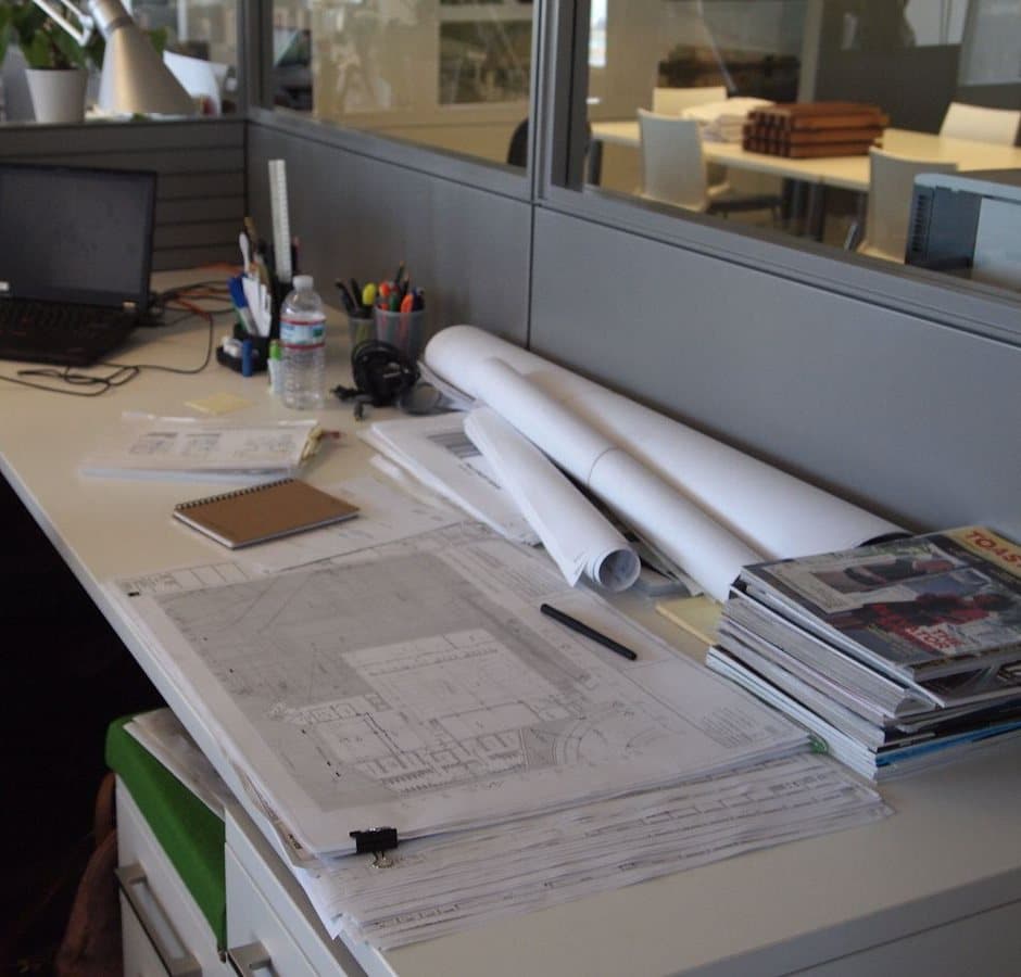 Plans and rolled drawings piled on an office desk next to a laptop